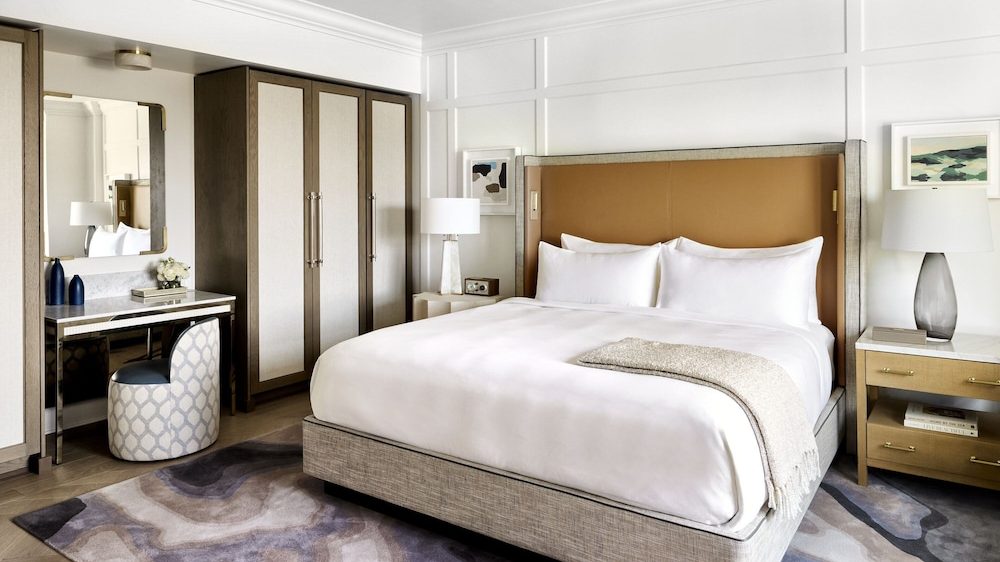 A modern bedroom featuring a large bed with a beige upholstered frame and a tan headboard, dressed in white bedding with a light beige throw blanket. On the right side, there is a wooden nightstand with two drawers, a table lamp with a gray glass base, and a small stack of books. On the left side, a white bedside table holds a white lamp with a clear base and a small clock. A wardrobe with dark wood and light beige panels stands next to a vanity desk with a mirror, a patterned round stool, and decorative items including two blue vases and a small flower arrangement. The floor has a patterned area rug in shades of gray, beige, and purple. The walls are white with subtle paneling, and two framed artworks hang above the nightstands.