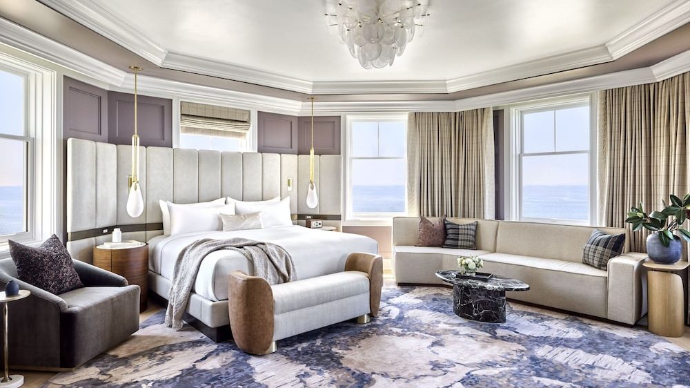 Spacious bedroom with a large upholstered bed featuring a tall, padded headboard and white bedding. A light gray bench with brown armrests is at the foot of the bed. To the left, there is a dark gray armchair with a patterned pillow and a round wooden side table. On the right side, a curved beige sofa with multiple pillows faces a black marble coffee table with a small flower arrangement. The room has large windows with beige curtains, light purple walls with paneling, and a patterned blue and beige carpet. A modern chandelier hangs from the ceiling.