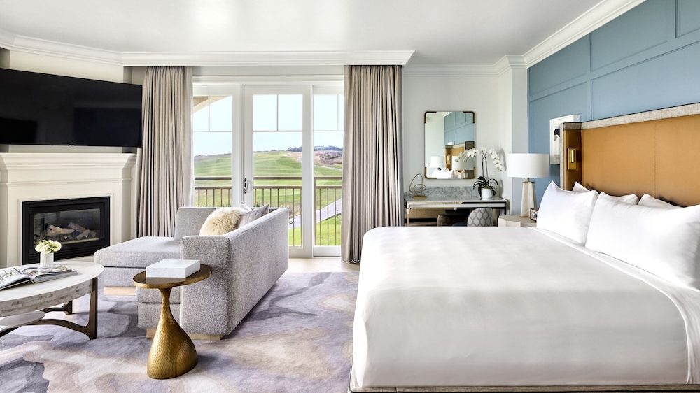 A modern hotel room featuring a large bed with white linens and a tan upholstered headboard against a blue accent wall. To the left, there is a gray sofa with a matching ottoman, positioned in front of a white fireplace and a mounted flat-screen TV. A round marble coffee table and a small gold side table are placed near the sofa. The room has large glass doors with beige curtains, offering a view of a green outdoor landscape. A vanity with a mirror, a lamp, and a white orchid plant is situated next to the bed. The floor is covered with a patterned carpet in soft neutral tones.