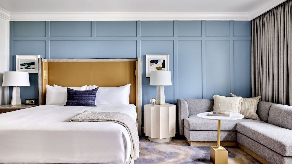 Modern bedroom with a large bed featuring white linens, a tan upholstered headboard, and a dark blue accent pillow. The wall behind the bed is painted blue with decorative paneling. On either side of the bed are white lamps on nightstands, with framed abstract art above. To the right, there is a gray L-shaped sofa with cream and beige pillows, a small white round table with a gold base, and gray curtains. The floor has a patterned rug in neutral tones.
