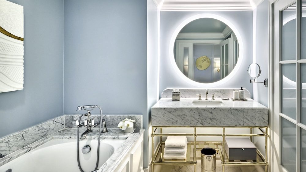 Bathroom with a marble countertop sink and matching marble bathtub surround. The sink has a gold metal frame base with white towels and a small trash bin underneath. A large round mirror with backlighting is mounted on the wall above the sink. The walls are painted light blue, and there is a small round magnifying mirror attached to the wall next to the sink. A glass door is visible on the right side.