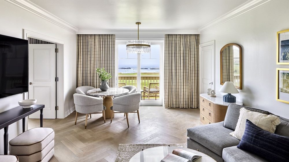 A modern living room with light wood herringbone flooring and white walls. A gray sectional sofa with cream and dark blue pillows sits on the right, next to a wooden dresser with a white marble top, a blue lamp, and a curved mirror above it. In the center, a round marble coffee table holds an open book. A round dining table with a marble top and four light gray upholstered chairs is positioned near large glass doors with beige plaid curtains, leading to a balcony with outdoor seating and a view of the ocean. A black console table with a white bowl and two beige ottomans are on the left, beneath a mounted flat-screen TV. A modern chandelier hangs above the dining table.