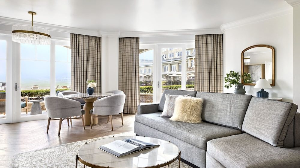 Living room with a large gray sectional sofa adorned with two decorative pillows, a round marble coffee table with an open book, and a dining area featuring a round table surrounded by four upholstered chairs. The space has large windows with plaid curtains, allowing natural light to fill the room, and a console table with a mirror, a plant, and a lamp against the wall.