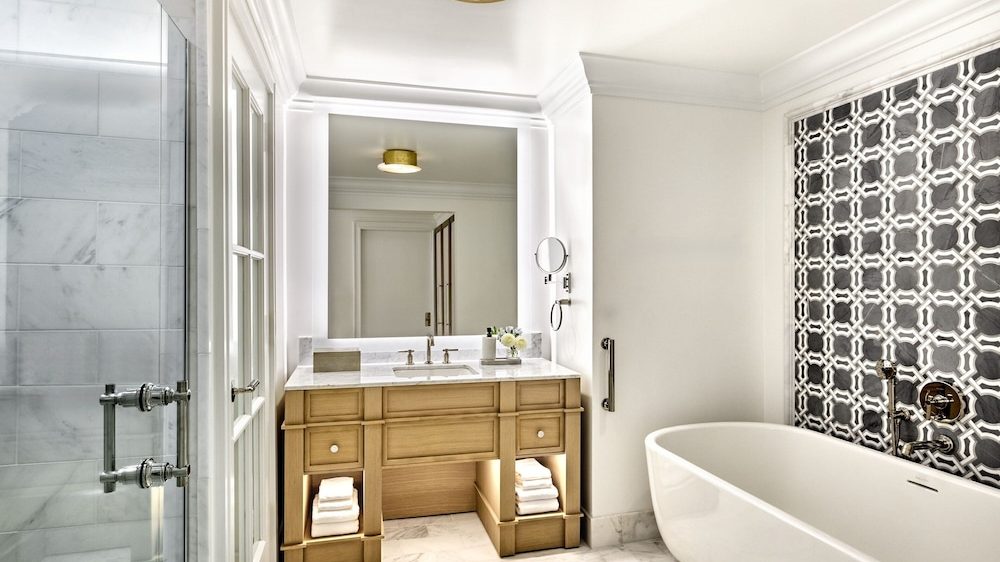 Modern bathroom featuring a wooden vanity with a white marble countertop and built-in sink, illuminated by a large backlit mirror. A freestanding white bathtub is positioned next to a wall with a decorative black and white geometric tile pattern. The floor is covered in white marble tiles, and a glass-enclosed shower is visible on the left side. The space is bright with white walls and ceiling, accented by a ceiling light fixture.