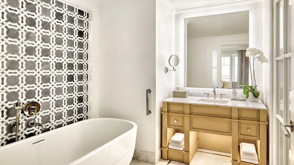 A modern bathroom featuring a white freestanding bathtub with a decorative black and white geometric tile wall behind it. Opposite the tub is a wooden vanity with a white marble countertop, an under-mounted sink, and a large backlit mirror. The vanity has open shelves with neatly folded white towels and a small potted orchid on the countertop. The floor is covered with light-colored marble tiles.