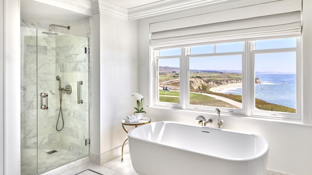 Modern bathroom featuring a white freestanding bathtub positioned under a large window with a scenic ocean and cliff view. To the left, there is a glass-enclosed shower with marble tile walls and a rainfall showerhead. A small round side table next to the bathtub holds a potted orchid and neatly folded towels. The room has white walls and marble flooring.