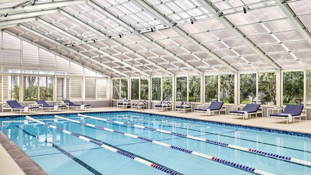 Indoor swimming pool with clear blue water, divided into lanes by floating lane markers. The pool area is enclosed by large glass windows and a transparent roof, allowing natural light to fill the space. Along the sides of the pool, there are several lounge chairs with dark cushions and small side tables. Trees and greenery are visible outside the windows.