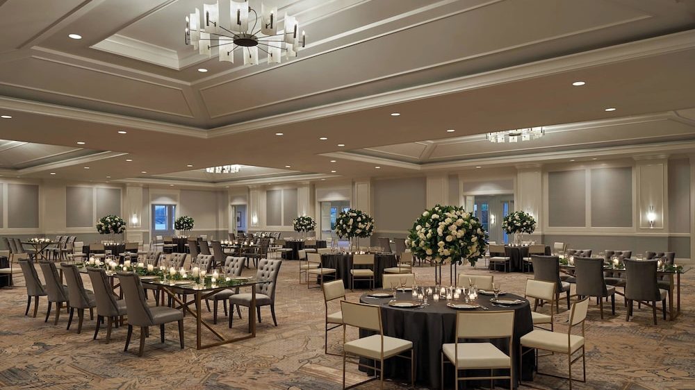 A spacious, elegant banquet hall with multiple round and rectangular tables set for an event. The tables are covered with black tablecloths and decorated with large floral centerpieces featuring white flowers and greenery. Each table has neatly arranged place settings and small candles. The room has a neutral color palette with beige walls, recessed lighting, and modern chandeliers hanging from the ceiling. The floor is covered with a patterned carpet in muted tones.