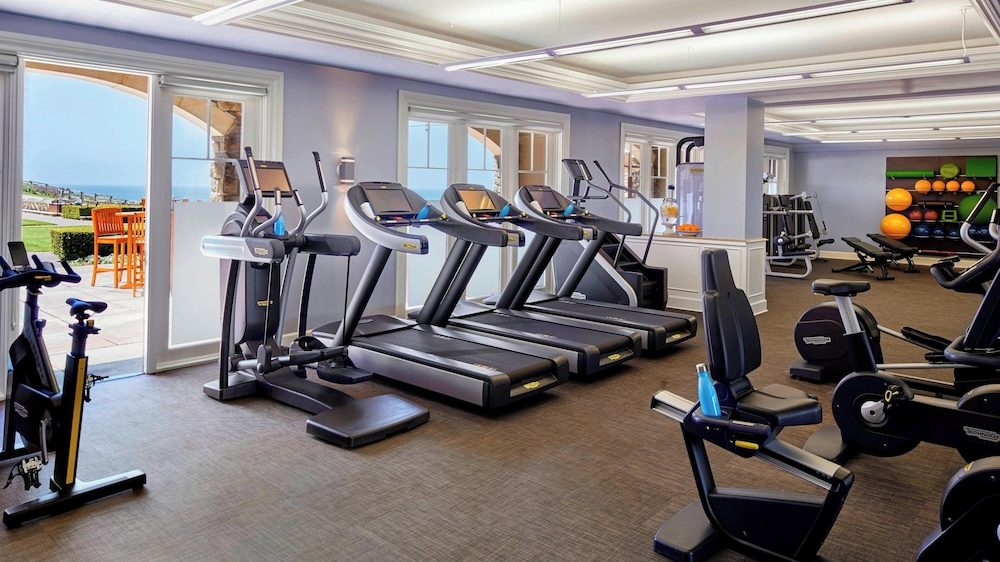A spacious gym with multiple treadmills, stationary bikes, and elliptical machines. The room has large windows and glass doors letting in natural light, with a view of an outdoor patio and greenery. There is a rack of various exercise balls and weights in the background. The floor is carpeted, and the ceiling has modern fluorescent lighting. A blue water bottle is placed on one of the exercise machines.