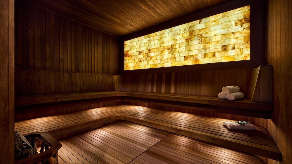 Wood-paneled sauna room with tiered wooden benches, soft under-bench lighting, a backlit rectangular salt wall panel, and neatly rolled white towels placed on the upper bench.