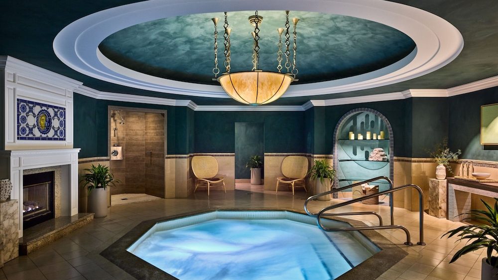 Indoor spa room featuring a lit octagonal hot tub with metal handrails, surrounded by beige tiled flooring. The walls are dark teal with a beige tile border, and the ceiling has a circular recessed design with a large hanging chandelier. There is a fireplace with decorative blue and white tile above it on the left, two wicker chairs against the back wall, a shower area with a glass door, and an arched alcove holding candles and towels. Potted plants and a countertop with a sink and decorative items are also visible.