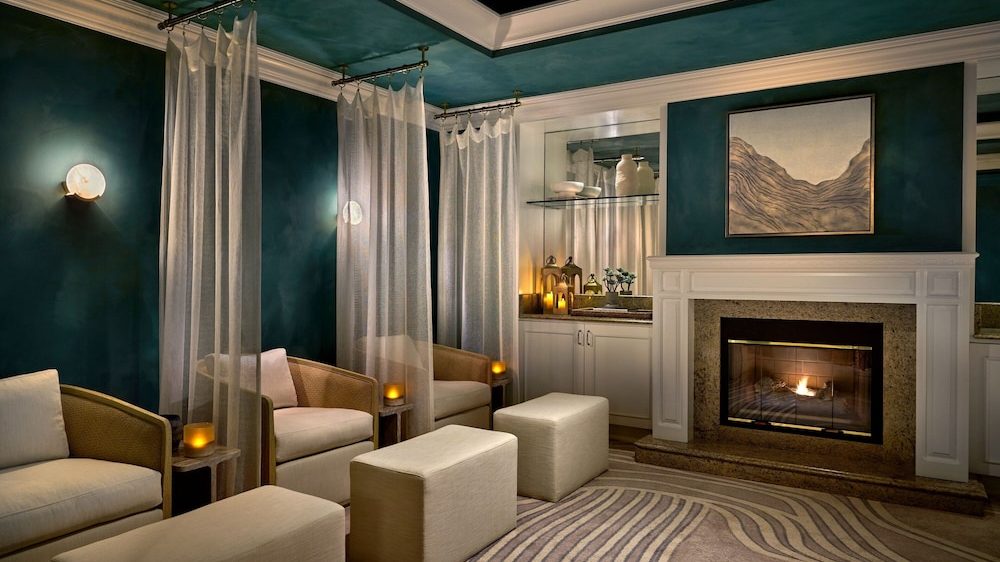 A cozy room with deep teal walls and ceiling, featuring three beige armchairs separated by sheer white curtains hanging from ceiling-mounted rods. Each chair has a small wooden side table with a lit candle. In front of the chairs are matching beige ottomans. A white fireplace with a granite surround is lit, and above it hangs a framed abstract artwork. To the right of the fireplace is a white cabinet with a mirrored backsplash, decorated with candles, vases, and small plants. The floor is covered with a beige and brown patterned rug.