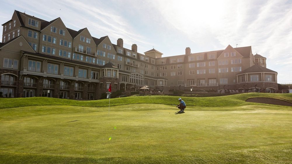 A large multi-story building with numerous windows and a stone and beige exterior overlooks a well-maintained golf green. A person wearing a blue shirt and cap is crouching near the hole with a red flag on the green, preparing to putt. The sky is partly cloudy with the sun shining brightly above the building.