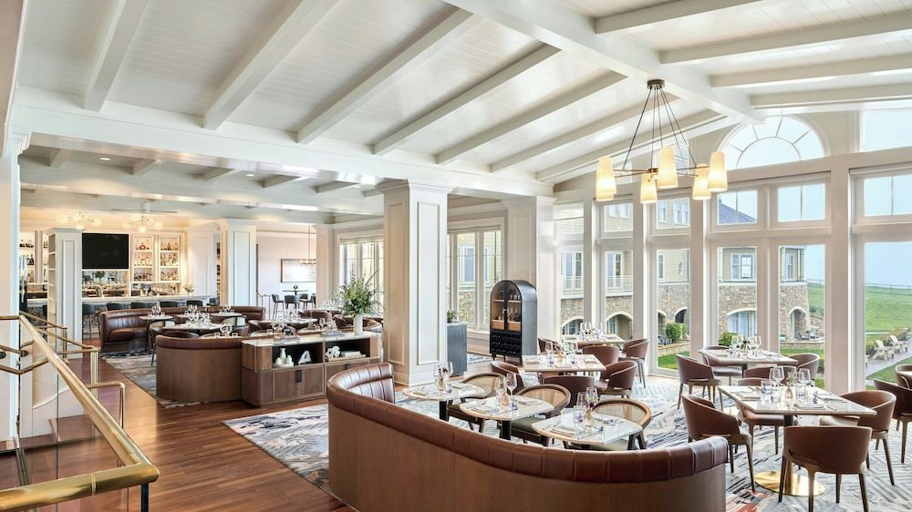 Spacious, elegant dining area with large windows offering a view of a stone building and green lawn outside. The room features white vaulted ceilings with exposed beams, wooden floors, and a mix of brown leather curved booths and wooden chairs around square tables set with glassware and napkins. A modern chandelier with multiple lights hangs from the ceiling, and a bar area with shelves of bottles and a TV is visible in the background.