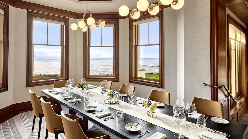 A modern dining room with a long black wooden table set for a meal. The table has a white runner, small floral centerpieces, white plates, folded napkins, and various glassware including wine glasses. Surrounding the table are tan leather chairs. The room features large windows with wooden frames, offering a view of the ocean and grassy area outside. Two contemporary chandeliers with round white bulbs hang from the ceiling. The walls are light-colored with wooden trim, and the floor has a striped carpet.