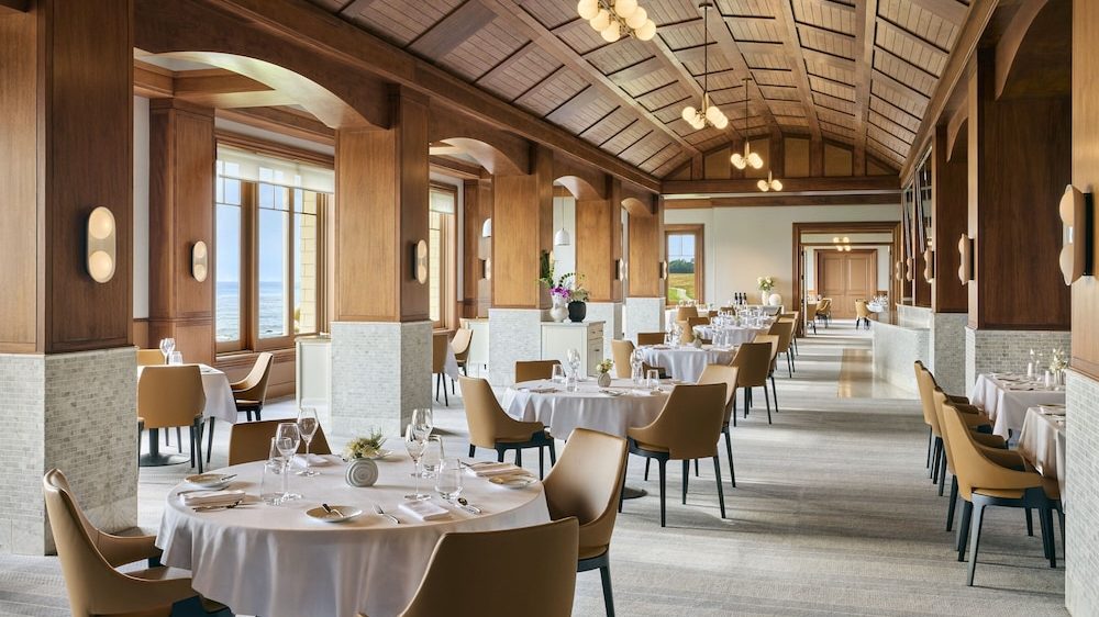 A spacious, elegant restaurant interior featuring round tables covered with white tablecloths, set with glassware, plates, and cutlery. The seating consists of tan upholstered chairs with black legs. The room has a high, wooden vaulted ceiling with modern pendant lights and wood-paneled columns with light-colored stone bases. Large windows on one side offer a view of the ocean, allowing natural light to fill the space. The overall design is warm and inviting with a mix of wood and neutral tones.