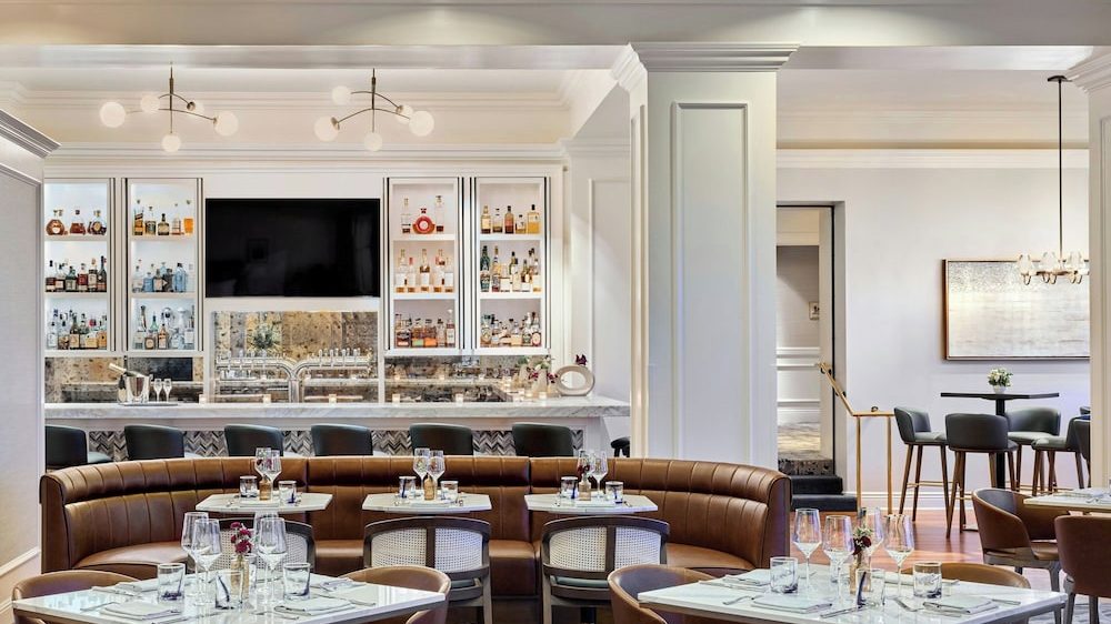 A modern bar and dining area featuring a curved brown leather banquette with small square tables set with glassware and cutlery. Behind the banquette is a marble-topped bar with a mirrored backsplash, glass-front cabinets displaying bottles of liquor, and a large flat-screen TV mounted in the center. The space is illuminated by contemporary pendant lights with round bulbs, and there are additional dining tables and chairs visible in the adjacent room. The overall decor is clean and elegant with white walls and ceiling beams.
