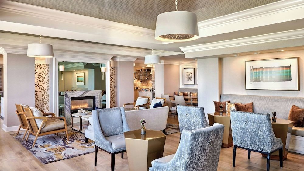 A modern, elegant lounge area with light wood flooring and a patterned ceiling. The space features a mix of seating including light blue upholstered chairs with ring handles on the back, beige cushioned armchairs with wooden frames, and a built-in bench with brown and beige throw pillows. There are small, gold-toned geometric tables with small flower vases on them. A fireplace with a marble surround is set into a mirrored wall, and abstract artwork is displayed on the walls. The room is softly lit by pendant lamps with white shades.