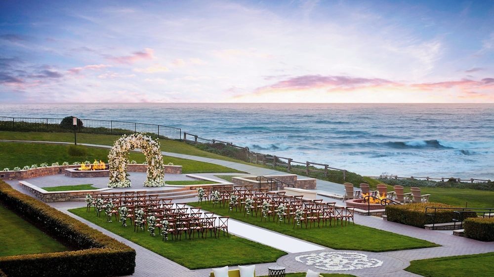 Outdoor wedding setup on a grassy area overlooking the ocean, featuring rows of wooden chairs with floral decorations, a flower-covered arch, a paved aisle, fire pits with seating, and a clear sky at sunset.