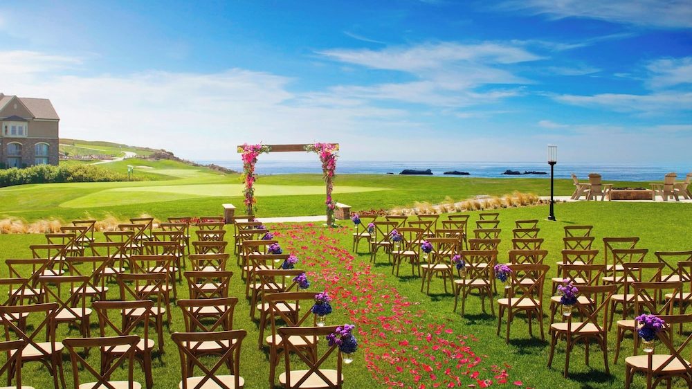 Outdoor wedding setup on a green lawn with wooden chairs arranged in rows on either side of an aisle covered with red flower petals. The aisle leads to a wooden arch decorated with pink flowers. In the background, there is a view of the ocean under a blue sky with scattered clouds. A house is visible on the left side, and some outdoor seating with a fire pit is on the right.