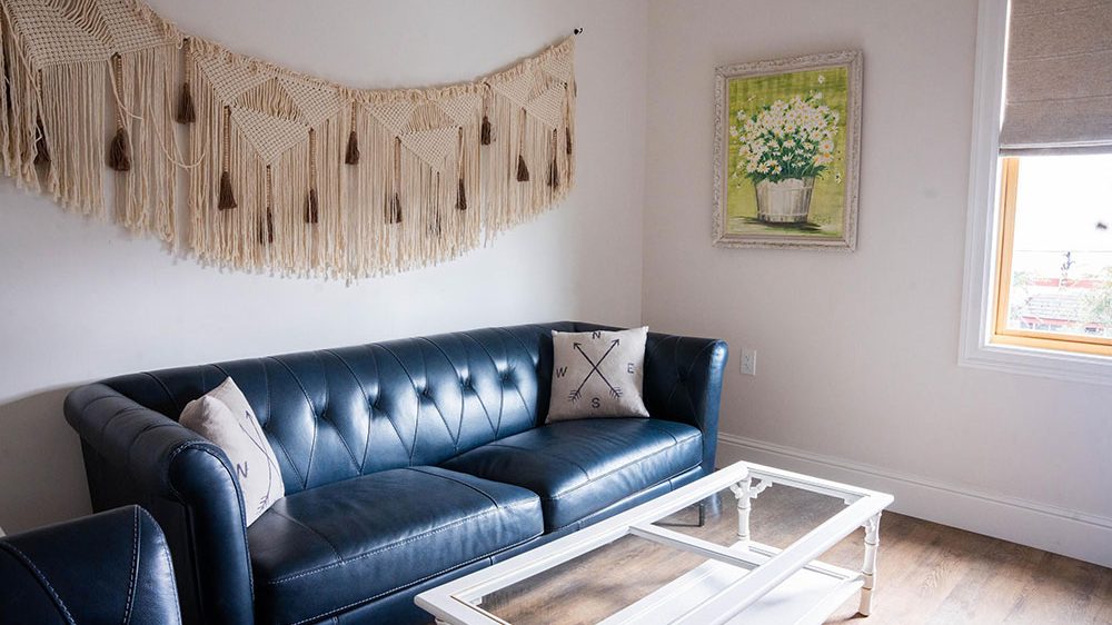 A modern living room features a dark blue tufted leather sofa with two decorative pillows, one with a compass design. Above the sofa hangs a beige macramé wall hanging with tassels. A white coffee table with a glass top is positioned in front of the sofa. On the adjacent wall, there is a framed painting of white flowers in a pot with a green background. A window with a beige Roman shade lets in natural light, illuminating the wooden floor.