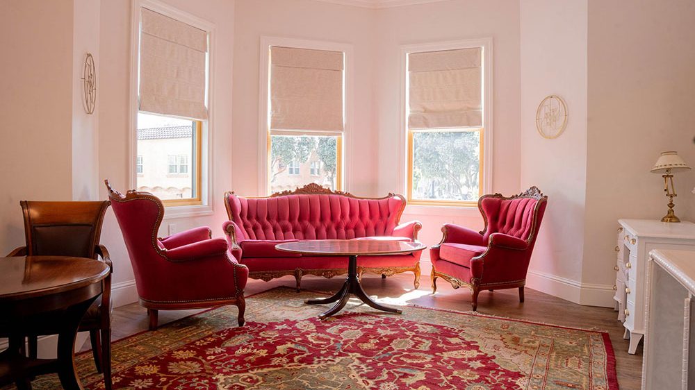 A cozy sitting area with a vintage-style red tufted sofa and two matching armchairs arranged around a dark wooden oval coffee table. The room features three tall windows with beige Roman shades, a large ornate red and beige patterned rug, light-colored walls, and a white dresser with a brass lamp on the right side.