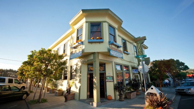 Two-story pale yellow building with green trim, featuring signs for "San Benito Restaurant & Saloon" and "SunButter Hostel." The building has flower boxes under the second-floor windows and a pumpkin near the entrance. Trees and parked cars line the sidewalk around the building.