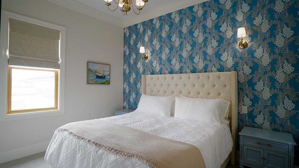 A bedroom with a large bed featuring a tufted beige headboard and white bedding with a beige throw blanket. The wall behind the bed is covered in blue wallpaper with a silver floral pattern. There are two brass wall sconces with white lampshades on either side of the bed. A brass chandelier with white lampshades hangs from the ceiling. On the right side of the bed is a light blue nightstand with two cabinet doors. A window with a beige Roman shade is on the left wall, and a small framed painting hangs between the window and the bed. The room has light-colored walls and carpeted flooring.