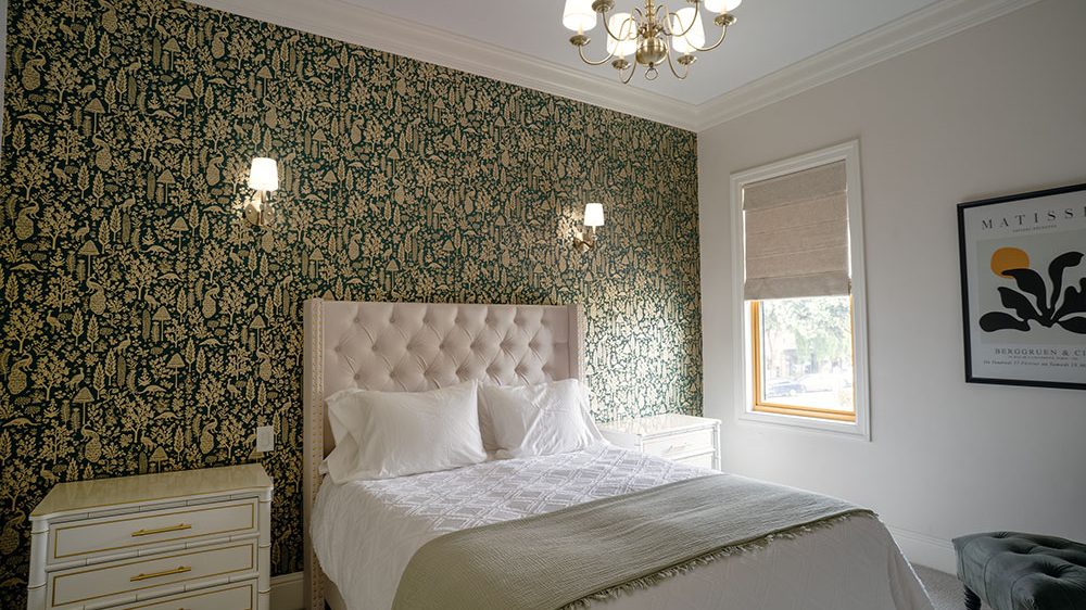 Bedroom with a tufted beige headboard bed dressed in white linens and a light green throw blanket. The wall behind the bed features dark green wallpaper with a gold botanical pattern. Two white nightstands with gold handles flank the bed. A window with a beige Roman shade is on the adjacent wall, next to a framed abstract art print. The room is lit by a chandelier and two wall sconces.