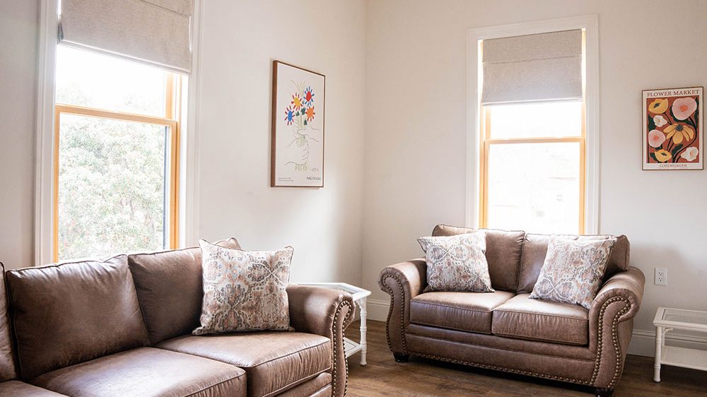 Two brown leather sofas with patterned throw pillows are arranged in a corner of a room with light-colored walls and wooden floors. Each sofa is positioned near a window with beige Roman shades. Between the sofas are white side tables, and two framed floral artworks hang on the walls above the sofas.
