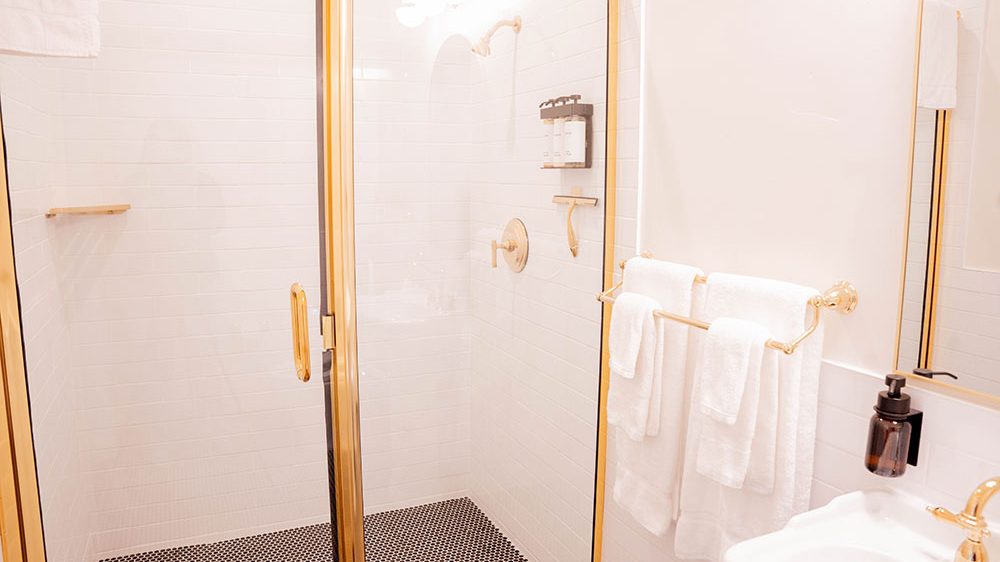 Shower area with gold-framed glass door, white tiled walls, and black hexagonal floor tiles. Inside the shower, there is a gold showerhead, a gold control knob, a wall-mounted dispenser with three bottles, and a gold squeegee holder. Outside the shower, a gold towel rack holds three white towels. A white sink with a gold faucet and a black soap dispenser is partially visible. The overall bathroom design features white and gold accents.