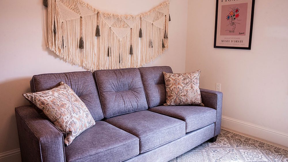 A gray upholstered three-seat sofa with two patterned throw pillows sits against a light-colored wall. Above the sofa is a large macrame wall hanging with tassels. To the right, a framed Picasso poster with a floral design hangs on the wall. The floor features a patterned rug in neutral tones.