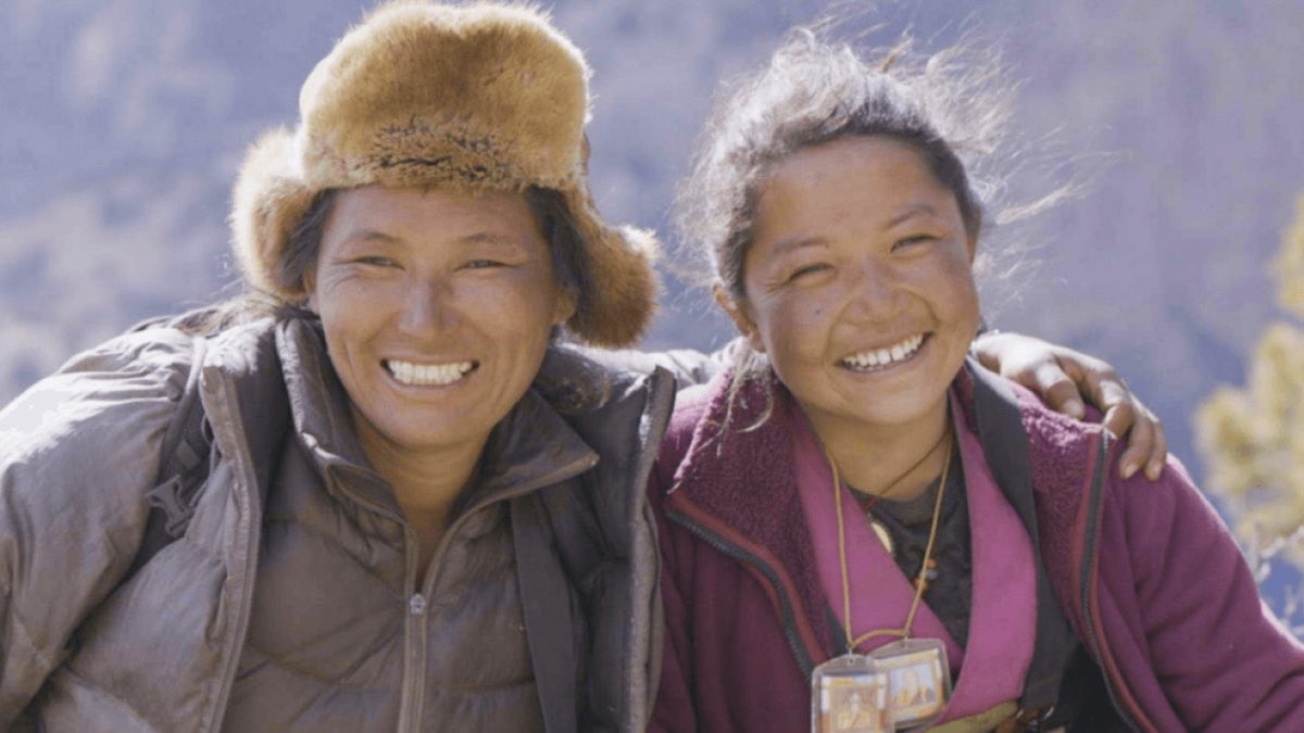 Two smiling people with arms around each other; the person on the left wears a brown fur hat and a gray jacket, while the person on the right wears a purple jacket and has a necklace with a small rectangular pendant.