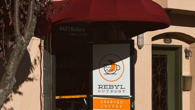 Entrance to a coffee shop with a maroon awning and a partially open door displaying the logo and text "REBYL OUTPOST," "GRAB+GO COFFEE," social media icons, and the website "REBYLFOOD.COM." A wooden bench is positioned to the left of the entrance, and a tree with dark leaves partially shades the area. The building exterior is beige with a green door numbered 725 visible to the right.