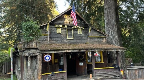 Rustic wooden building labeled “APPLE JACK’S” with an American flag, surrounded by tall trees and a motorcycle in front.