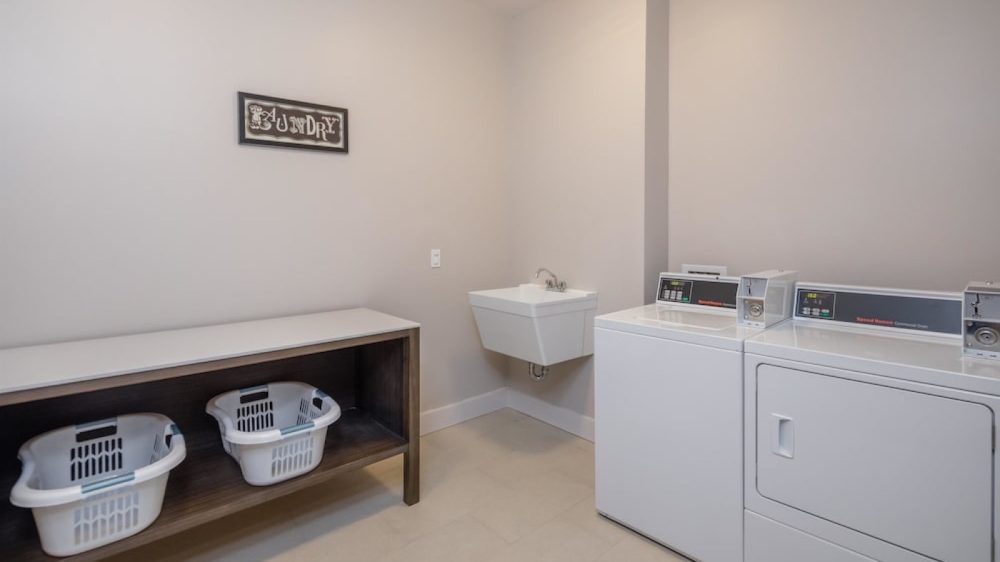Laundry room with a white washing machine and dryer, a wall-mounted utility sink, a wooden table with two white laundry baskets underneath, and a framed "Laundry" sign on the wall. The walls are light beige and the floor is tiled.