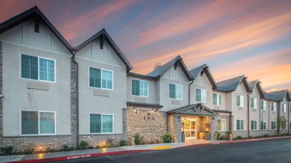 A two-story hotel building with a beige exterior and stone accents, featuring multiple windows with white shutters. The entrance has a covered porch with wooden beams and warm lighting. The hotel name "Aristocrat Hotel" is displayed on the stone wall near the entrance. The sky above shows a colorful sunset with shades of pink, orange, and blue.