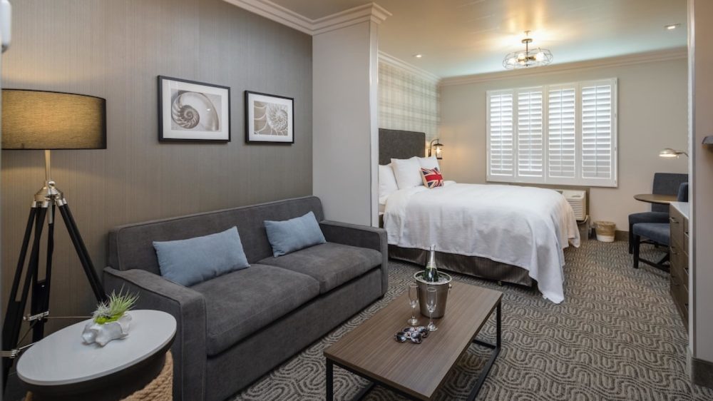 Modern hotel room with a gray sofa featuring two light blue pillows, a wooden coffee table with a champagne bottle in an ice bucket, two champagne glasses, and sunglasses. The bed has white linens and a pillow with a Union Jack design. The room includes a tripod floor lamp, two framed shell prints on the wall, a small round table with two chairs, and a window with white plantation shutters. The carpet has a geometric pattern, and the walls are painted in neutral tones with crown molding.
