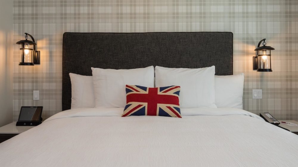 King-sized bed with a dark gray upholstered headboard, white bedding, and four white pillows. A small rectangular pillow with a Union Jack design is centered in front of the white pillows. The wall behind the bed has a light plaid pattern. Two black lantern-style wall lamps are mounted on either side of the headboard, above white nightstands. One nightstand has a digital device, and the other has a telephone and a notepad with a pen.