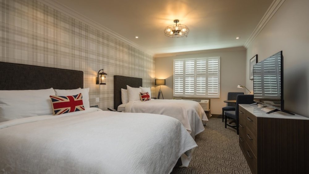 Two queen beds with white bedding and dark upholstered headboards in a hotel room. Each bed has a pillow with a Union Jack design. The room features a plaid accent wall behind the beds, a patterned carpet, a window with white plantation shutters, a floor lamp, a flat-screen TV on a wooden dresser, and a small table with two chairs. The ceiling has a modern light fixture.