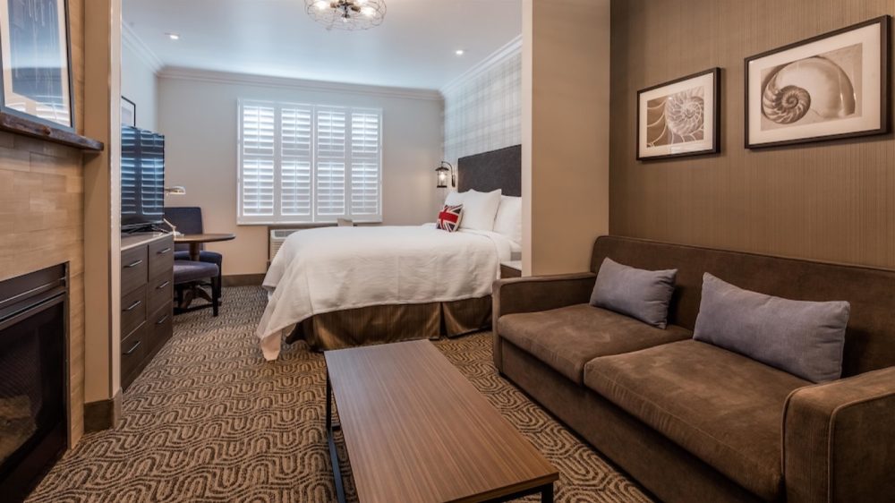 Hotel room with a bed featuring white linens and a pillow with a Union Jack design, a brown sofa with two gray cushions, a wooden coffee table, a patterned carpet, a TV on a wooden dresser, a small round table with two chairs, and two framed shell prints on the wall.