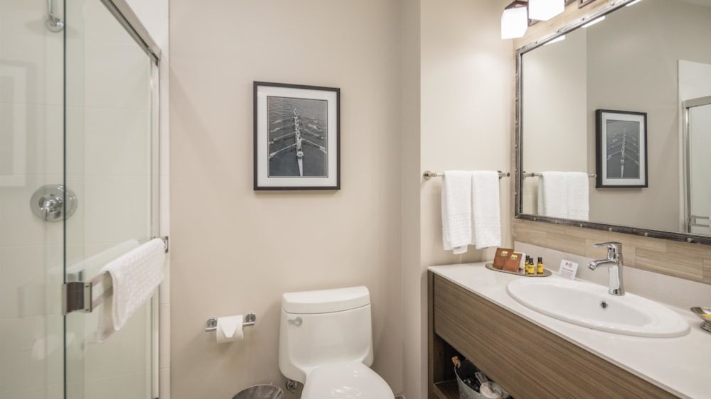 Modern bathroom with a white toilet, glass shower door with a towel hanging on the handle, and a wooden vanity with a white sink. Above the sink is a large mirror with four square light fixtures. A towel rack with two white towels is mounted on the wall next to the vanity. A framed black-and-white photo hangs on the wall above the toilet. A small trash can is placed beside the toilet. The overall color scheme is neutral with beige walls and light wood tones.