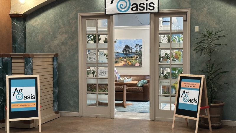 Entrance to an art studio named "Art Oasis" with double glass doors. Above the doors is a white sign with the studio's name and a blue spiral design. On either side of the entrance are sandwich boards displaying the "Art Oasis" logo and welcoming messages. Inside, there is a brown leather couch, a wooden table, plants, and a colorful painting of a landscape with trees and flowers on the back wall. The walls around the entrance are textured green with beige trim, and there is a potted plant to the right of the doors.