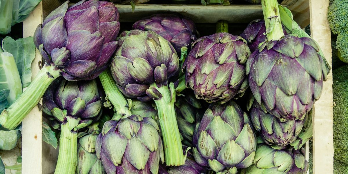 A wooden crate filled with fresh artichokes, displaying a mix of purple and green hues on their thick, layered leaves and long green stems. Some broccoli and cabbage leaves are partially visible around the crate edges.