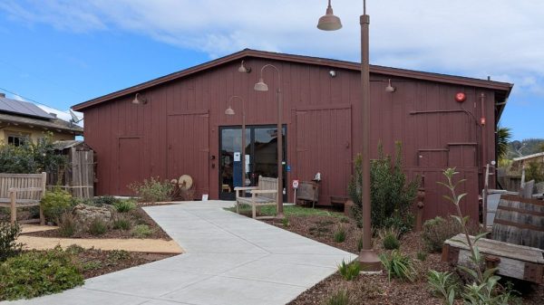 A brown wooden building with a peaked roof and a glass door entrance. A concrete walkway leads up to the door, flanked by garden beds with shrubs and plants. There are several brown street lamps along the walkway and wooden benches on either side near the entrance. The sky is partly cloudy.