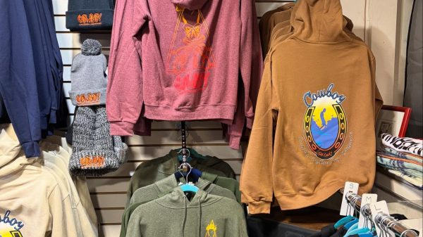 Display of various hoodies and beanies in a retail setting. Hoodies come in colors including maroon, mustard, cream, olive green, and black, with some featuring "Cowboy" and horseshoe designs. Beanies in black and gray have "Cowboy" embroidered in orange. Above the display is a "Salty Crew" sign, and two framed photos of surfers riding waves are mounted on the wall.
