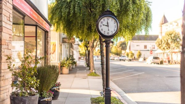 Black vintage street clock on a sidewalk under a leafy tree, showing the time as 11:45. The sidewalk is lined with potted plants and a building with a red sign advertising local and imported wines and a wine tasting bar. The street is quiet with a few parked cars and buildings in the background.