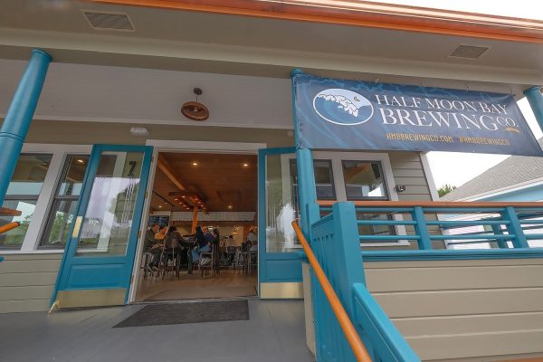 Entrance to Half Moon Bay Brewing Co. with open blue double doors, blue railings, and a banner displaying the brewery's name and website. Inside, people are seated at the bar.
