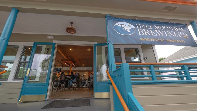 Entrance to Half Moon Bay Brewing Co. with open blue double doors, blue railings, and a banner displaying the brewery's name and website. Inside, people are seated at the bar.