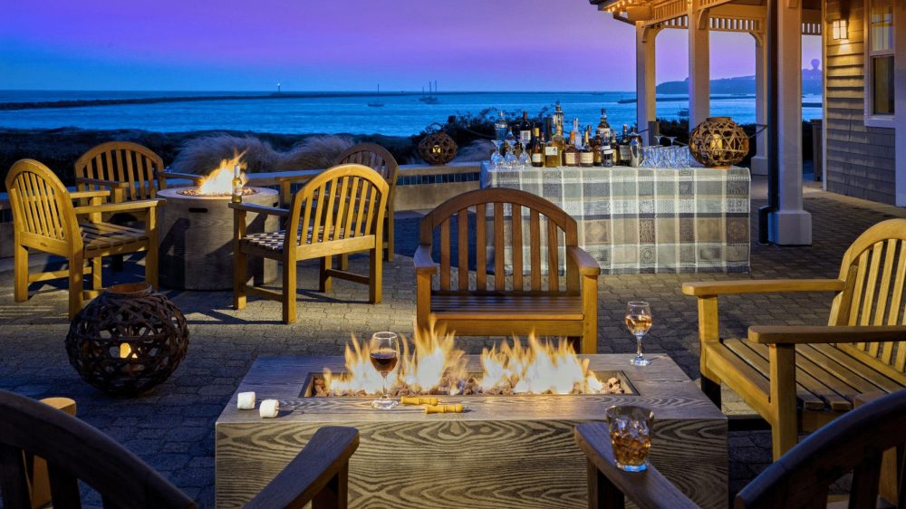 Outdoor patio seating area at dusk with wooden chairs arranged around two fire pits, one rectangular and one circular. The rectangular fire pit has flames and two glasses of wine on its edge, along with marshmallows and skewers. In the background, a table covered with a patterned cloth holds various bottles of liquor and glassware. The scene overlooks a calm body of water with boats visible in the distance under a purple-blue sky. Warm string lights adorn the porch of the nearby building.